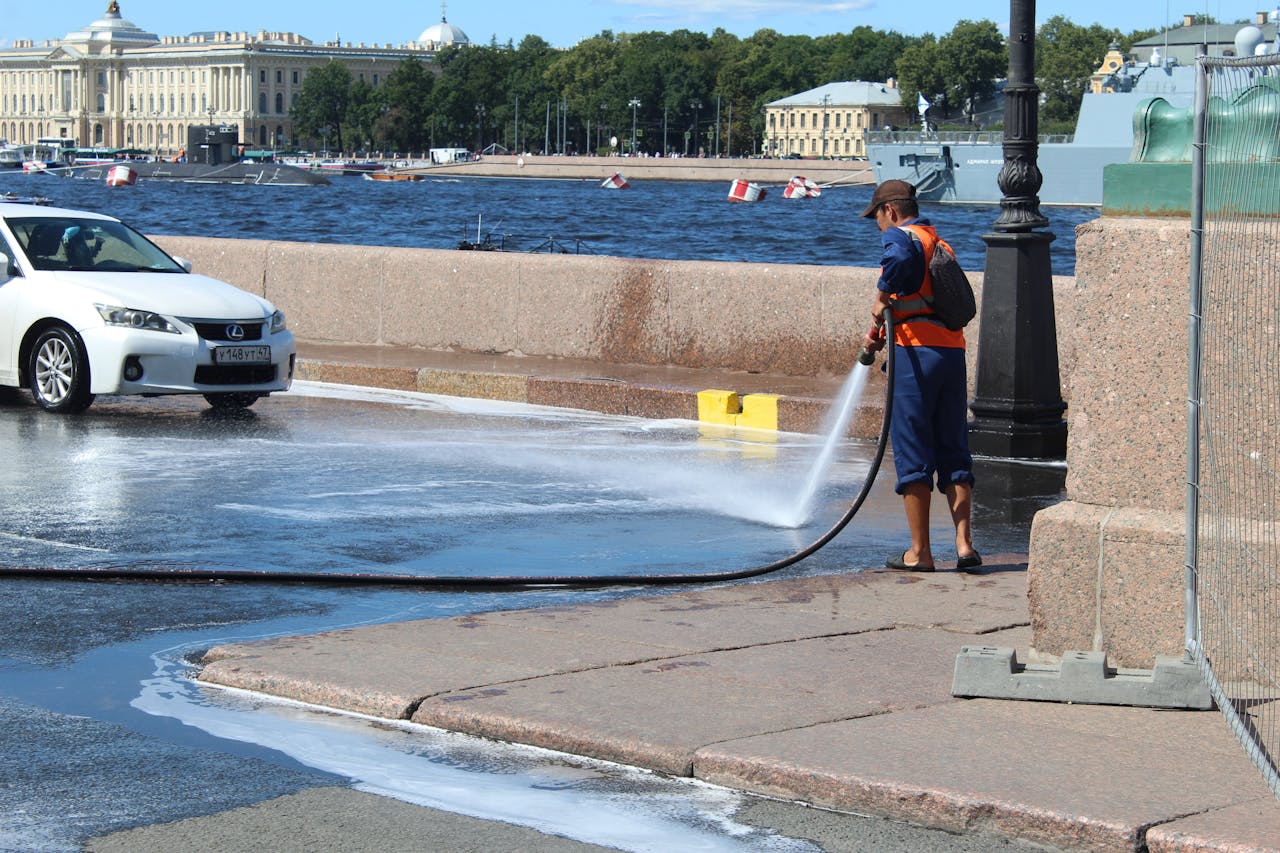 about-01 A street cleaner uses a hose to wash a sidewalk near the river, with a cityscape in the background.
